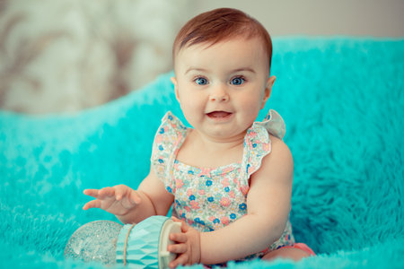 Cute baby with blue eyes and floral dress smiling and sitting on her bedの写真素材