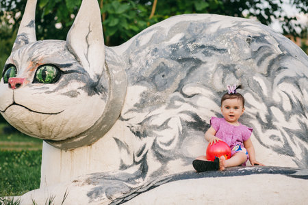 Playful baby with purple dress and butterfly bow holding red ball and sitting on stone cat statueの写真素材