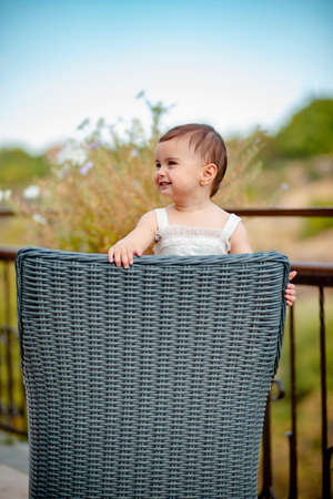 Vertical shot of adorable baby girl with white dress standing on straw chair and laughingの写真素材