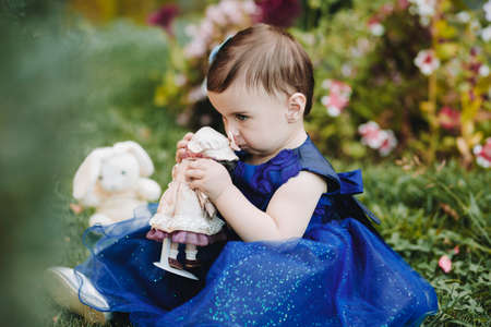 Cute baby girl holding and touching her toy with nose and sitting on grass in gardenの写真素材