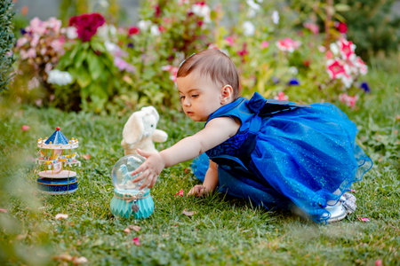 Princess baby girl with blue dress sitting on grass in garden and playing with one of her toysの写真素材
