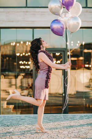 Cheerful girl with purple dress holding balloons raising her leg standing in front of restaurant and celebrating her birthdayの写真素材