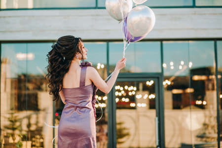 Closeup of luxury girl with purple dress celebrating her birthday, standing in front of restaurant with glass doors and holding her birthday balloonsの写真素材