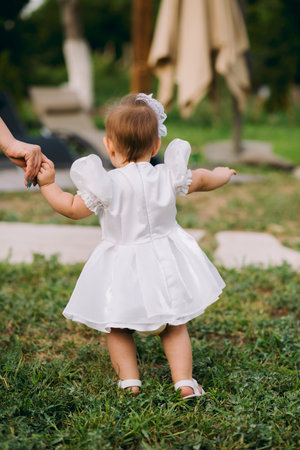 Adorable baby girl in white dress walking outdoors on grass with her mother holding her handの写真素材