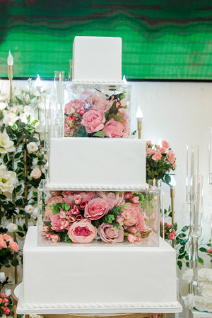 Sweet white engagement cake in banquet hall decorated with artificial flowers and lots of roses behindの写真素材