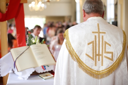 Bride and groom during a wedding ceremony in the background and the priest i the foreground