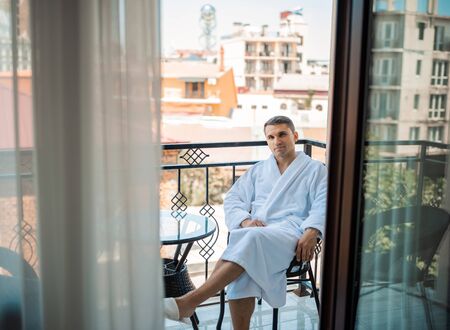 Handsome bearded man in the morning sitting in a hotel room in a robe. Young man wearing white bathrobe after shower bath. Gygiene conceptの写真素材