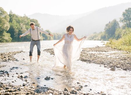 happy, emotional gorgeous bride and stylish groom having fun, splashed with water. Wedding couple running along the mountain river with bare feet.の写真素材