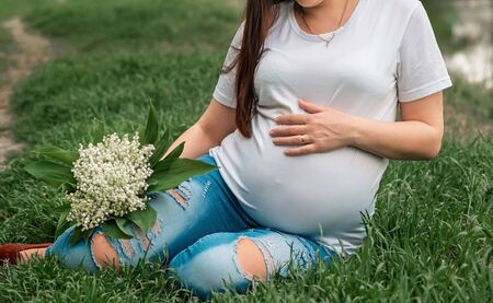 Closeup of pregnant woman, wearing white T-shirt, holding in hands bouquet of lilies of the valley outside. Pregnant woman holding her tummy.の写真素材