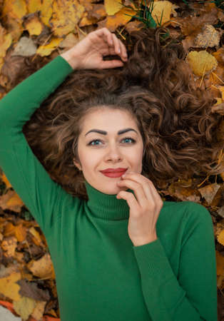 Curly cute girl with open eyes liying in the autumn leaves, holding hand near face and smile. Female...