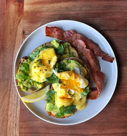 Eggs Benedict. Top view. Poached egg on white toasted bread with smashed avocado, rocket, bacon, Hollandaise sauce and lemon on a gray plate. Morning breakfast.の写真素材