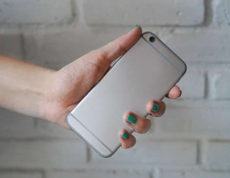 Gray smart phone in female hand with blue nails over rustic background, back view. Mockup for stickerの写真素材