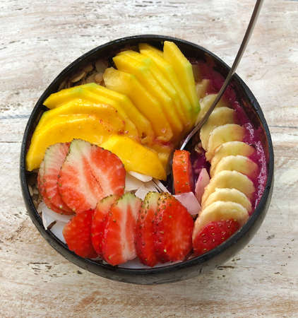 Pitaya breakfast bowl. Acai superfood smoothie bowl with mango, strawberry, banana, dragon fruit, granola and coconut flakes. White rustic wooden background. Overhead, top view, flat lay, copy space.の写真素材