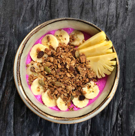 Pitaya breakfast bowl. White rustic black wooden background. Acai superfood smoothie bowl with pineapple slices, banana, dragon fruit smoothie, granola. Overhead, top view, flat lay, copy space.の写真素材