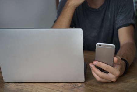 Closeup shot of a young man typing a text on mobile phone. Guy holding a modern smartphone and writing a phone message with laptop on table.の写真素材