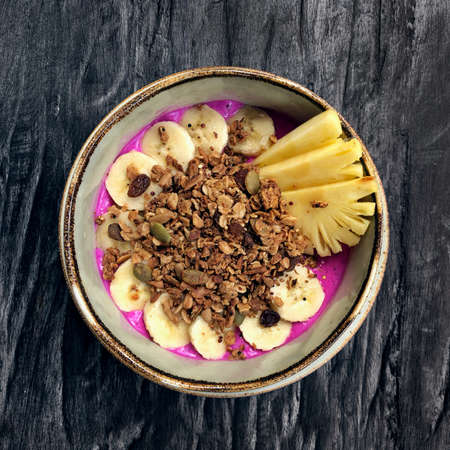 Pitaya breakfast bowl. White rustic black wooden background. Acai superfood smoothie bowl with pineapple slices, banana, dragon fruit smoothie, granola. Overhead, top view, flat lay, copy space.の写真素材