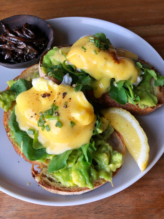 Eggs Benedict. Top view. Poached egg on white toasted bread with smashed avocado, rocket, bacon, Hollandaise sauce and lemon on a gray plate. Morning breakfast.の写真素材