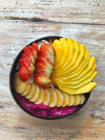 Pitaya breakfast bowl. Acai superfood smoothie bowl with mango, strawberry, banana, dragon fruit, granola and coconut flakes. White rustic wooden background. Overhead, top view, flat lay, copy space.の写真素材