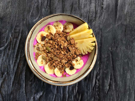Pitaya breakfast bowl. White rustic black wooden background. Acai superfood smoothie bowl with pineapple slices, banana, dragon fruit smoothie, granola. Overhead, top view, flat lay, copy space.の写真素材
