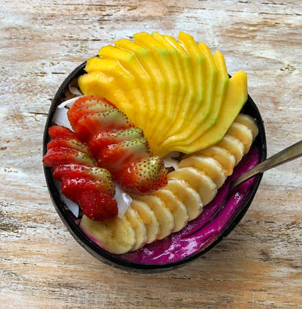 Pitaya breakfast bowl. Acai superfood smoothie bowl with mango, strawberry, banana, dragon fruit, granola and coconut flakes. White rustic wooden background. Overhead, top view, flat lay, copy space.の写真素材