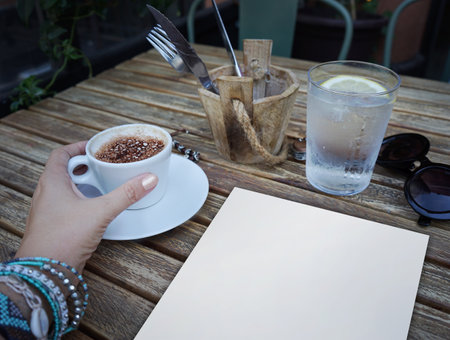 Female hand holding cup of coffee. Cafe wooden table with a glass of water, sunglasses. Rustic texture.の写真素材
