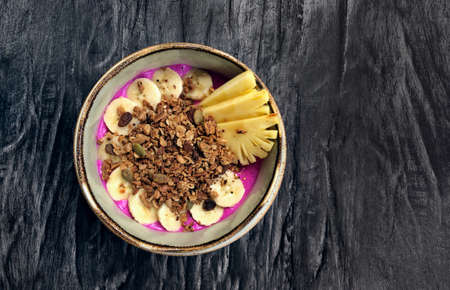 Pitaya breakfast bowl. White rustic black wooden background. Acai superfood smoothie bowl with pineapple slices, banana, dragon fruit smoothie, granola. Overhead, top view, flat lay, copy space.の写真素材
