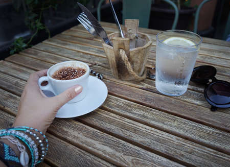 Female hand holding cup of coffee. Cafe wooden table with a glass of water, sunglasses. Rustic texture.の写真素材