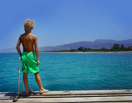 Island summer holidays: Handsome Caucasian boy standing on the dock and looking at blue sea.の写真素材