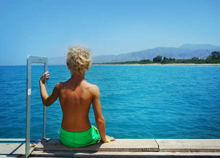 Island summer holidays: Handsome Caucasian boy standing on the dock and looking at blue sea.の写真素材