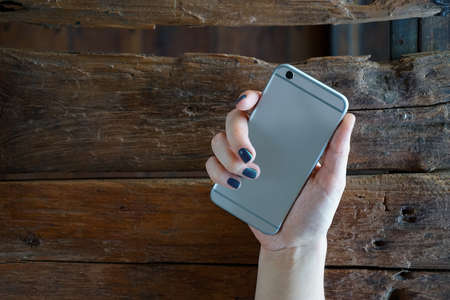 Gray smart phone in female hand with blue nails over rustic background, back view. Mockup for stickerの写真素材