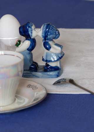 Kissing boy and girl. blue and white statue stands next to a pair of tea, boiled egg and spoon on a blue tablecloth and white napkinの写真素材