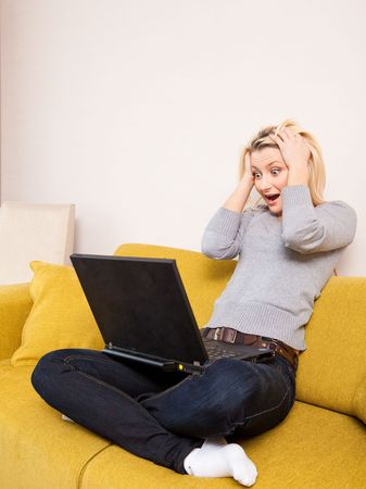 surprised young blond woman sitting on couch and looking at computerの写真素材