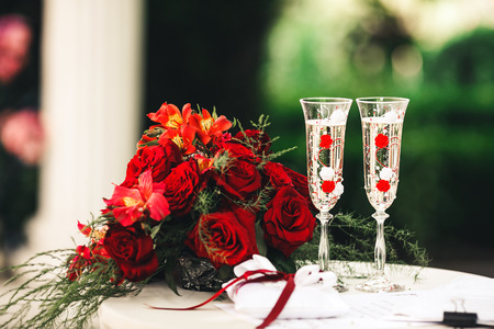 A red bouquet, beautiful glasses with champagne and a pillow for wedding rings lie on the table.の写真素材