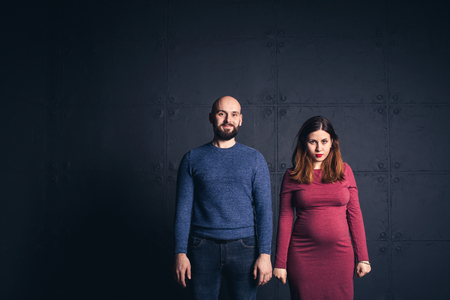 bearded man and pregnant woman looking in camera in studio on dark background. Copy spaceの写真素材