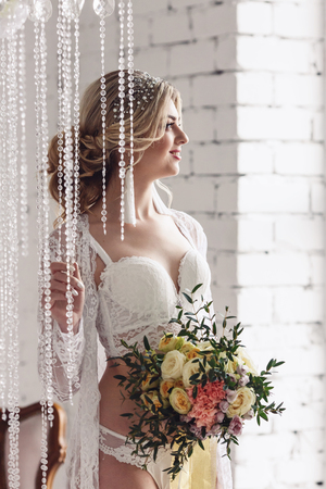 beautiful girl dressed in peignoir and underwear, stands near crystal curtains and holds bouquet in her handsの写真素材