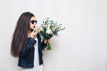 Beautiful stylish girl in sunglasses, dressed in leather jacket, holding bouquet of flowers and posing on white wallの写真素材