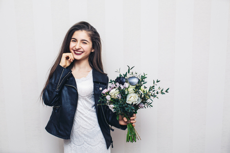 Beautiful stylish girl dressed in leather jacket, holding bouquet of flowers and posing against white wallの写真素材