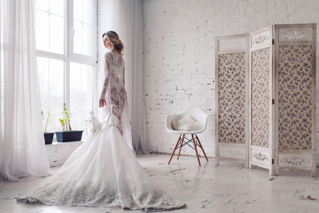 bride, dressed in peignoir and underwear, stands near window and holds wedding dress in her hands. Morning of brideの写真素材