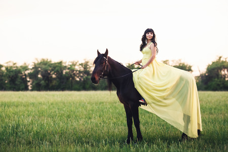 Beautiful girl in yellow dress sits on horse on field.の写真素材