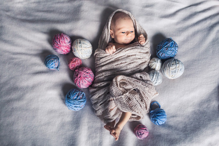 Baby in blanket lies on bed among tangles of yarn.