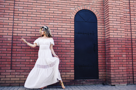 Beautiful girl in white dress is standing near brick wall with metal door and posing.の写真素材