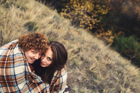 Loving cute couple sitting on grass and hiding behind rug in autumnの写真素材