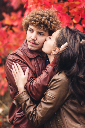 Curly-haired mustachioed man and brown-haired woman hugging in autumn against background of red trees. Guy passionately strokes girl's hair. Happy couple in loveの写真素材