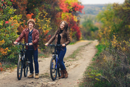 curly-haired mustachioed guy and an ore-haired girl in autumn stand with bicycles on road and show cheerful emotionsの写真素材