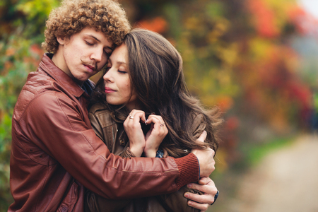 happy couple in love in autumn. curly-haired mustachioed man and ruddy-haired woman in leather jackets and jeans tenderly embrace. girl makes palms of heartの写真素材