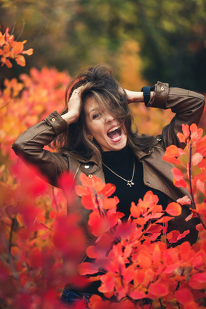 Cheerful happy ruddy-haired girl opens her mouth wide and ruffles her hair autumn between red bushes