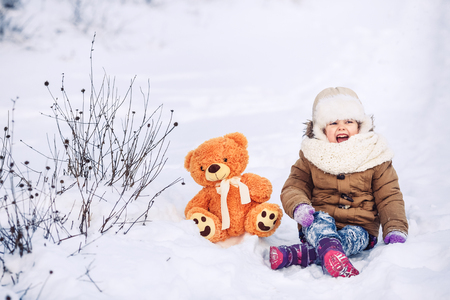 A cheerful little girl is sitting in the snow with her teddy bear. The girl laughs very much with her mouth open.の写真素材