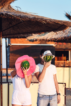 A guy and a girl are standing on the beach and shielding their heads with watermelons in hatsの写真素材