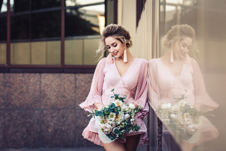 attractive young girl in a short dress with a flower bouquet posing on the street near the building wallの写真素材