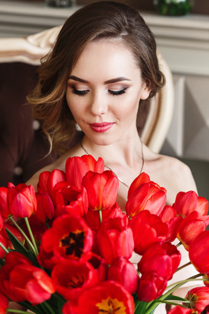 A beautiful young girl is sitting on the sofa and holding a large bouquet of red tulips. March 8 concept. Morning of the brideの写真素材